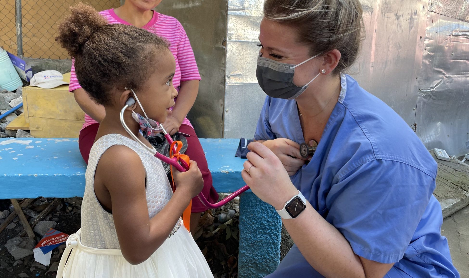 Doctor in a mask and scrubs letting a little girl test out their stethoscope